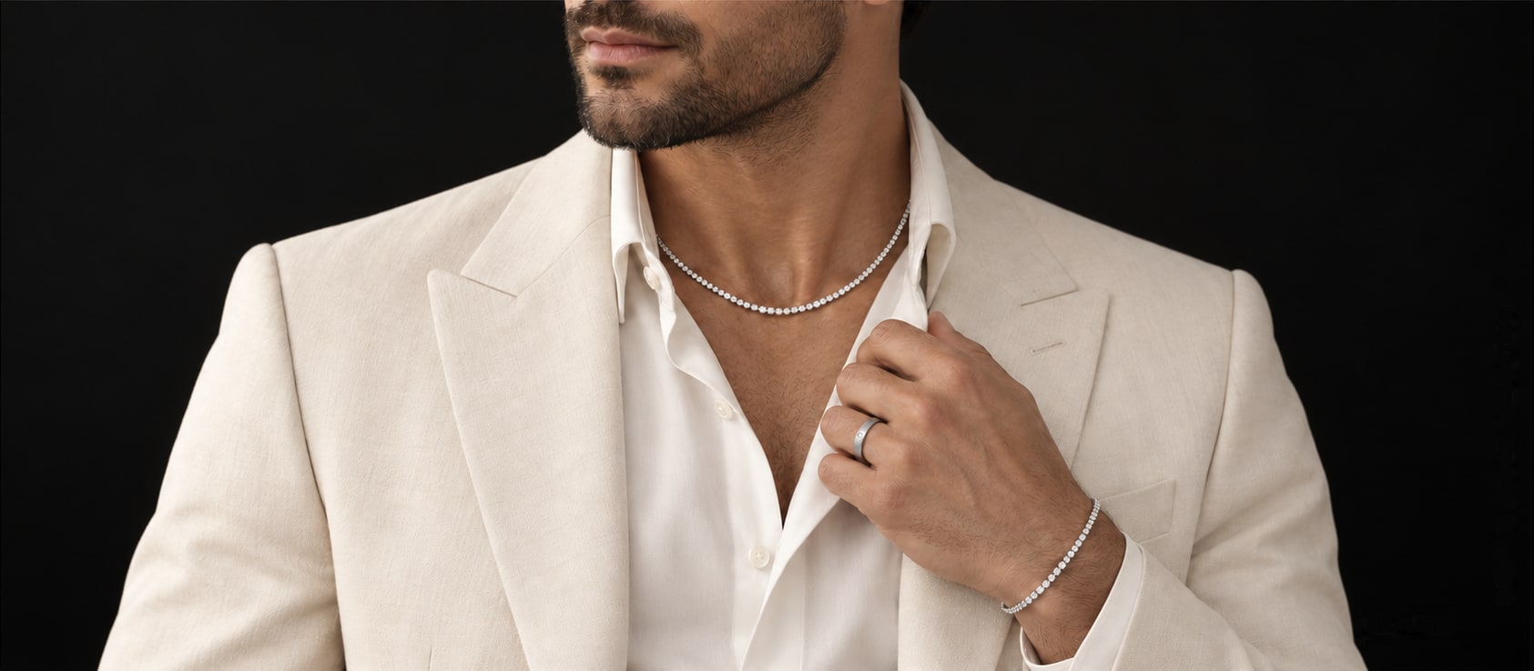 Man in a cream blazer and white shirt wearing a slim diamond tennis necklace, a diamond tennis bracelet and a plain band ring, on a black backdrop